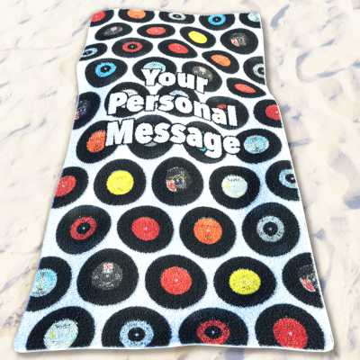 A beach towel lying on a beach, the towel having a mosaic of vinyl records on it along with a personal message