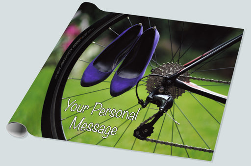 A roll of wrapping paper showing the rear wheel of a racing bicycle with a pair of purple heels hung from the spokes of the wheel, there is grass and trees in the background, and a personal message