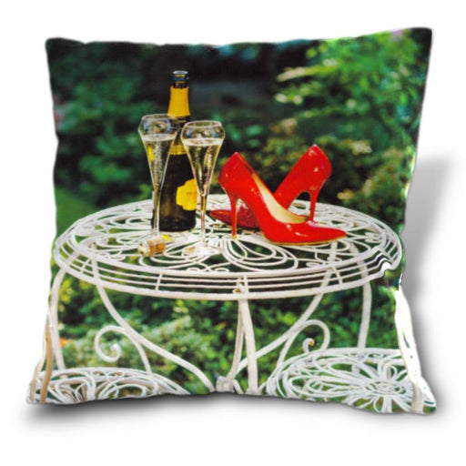 A cushion, the cushion having an image of a pair of red shoes on a white metal garden table, sat next to a bottle of fizzy wine and two poured glasses