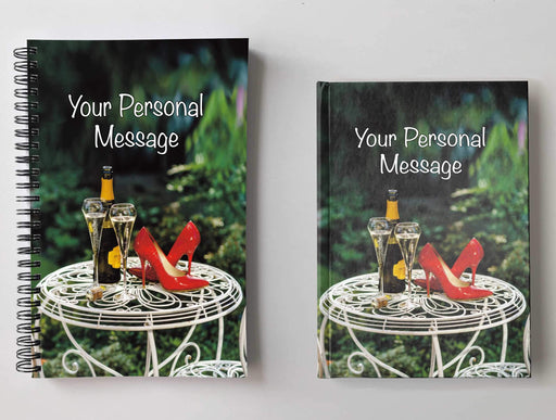 Two notebooks, side by side, both having covers with an image of a pair of red high heel shoes on a garden table adjacent to a bottle of sparkling wine and two filled glasses, and a personal message on the cover