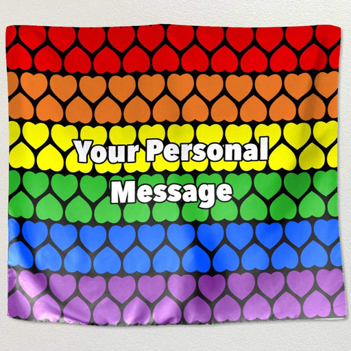 A tapestry hung on a white wall, the tapestry has horizontal rows of hearts, each row a differnet rainbow colour, and there is a lpersonal message printed