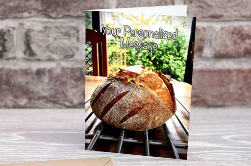a birthday or greetings card on a desk with an envelope and pen, the card having an image of a sourdough loaf of bread sat on a table with a view of a garden in the distance