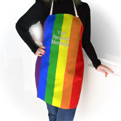 A woman wearing an apron, the apron has vertical stripes patterned from the pride rainbow flag, the woman has a black top and blue jeans and is leaning against a wall, the wall being very faded and faint