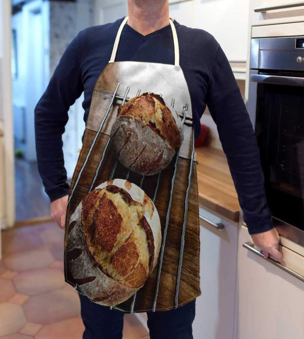 A kitchen apron being worn by a man in a kitchen, the apron having an image of a large sourdough loaf two sourdough loaves seen from an angled position