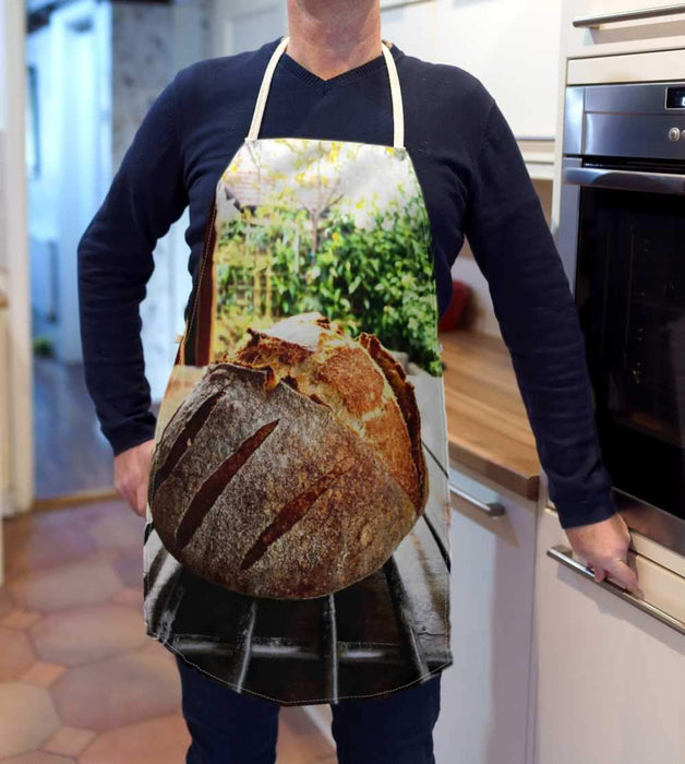 A kitchen apron being worn by a man in a kitchen, the apron having an image of a large sourdough loaf with a view of the garden in the rear of the image