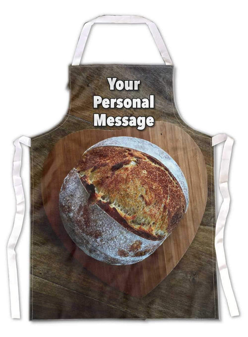 A kitchen apron flat on the floor, the apron having an image of a large sourdough loaf on top of a wooden heart shaped tray