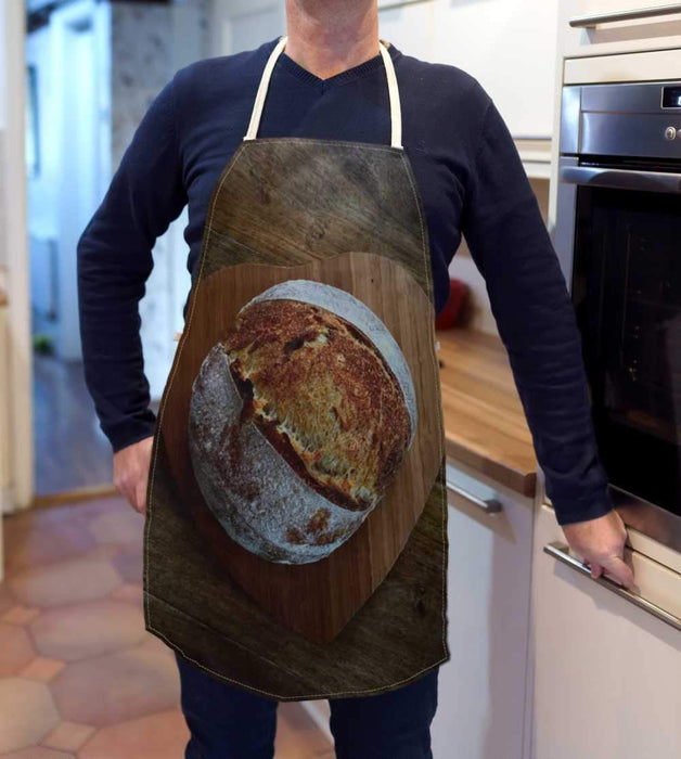 A kitchen apron being worn by a man in a kitchen, the apron having an image of a large sourdough loaf on top of a wooden heart shaped tray