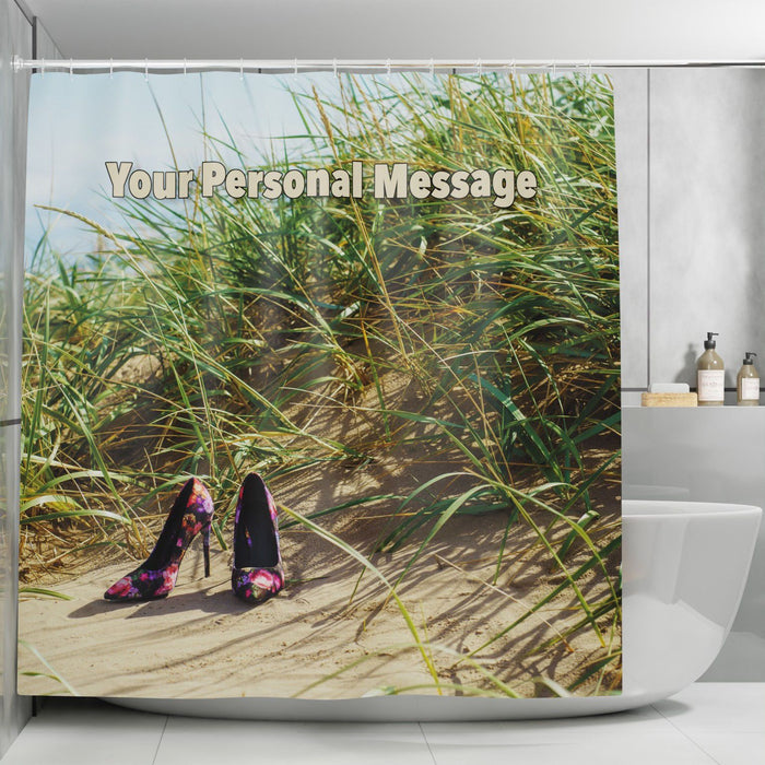 A shower curtain in a bathroom, the shower curtain having an image of a pair of high heel shoes on the sand at a beach near to some grassy sandhills on a sunny day, along with a personal message printed on the curtain