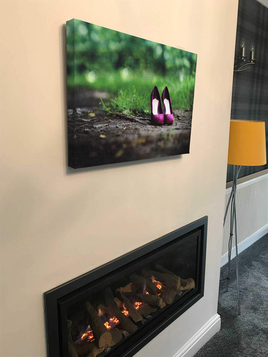 A canvas print of a pair of purple high heel shoes on a path within the woods, the canvas print is hung upon a wall in a house above a fire
