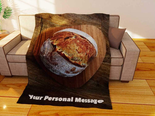 a fleece blanket flat on the couch, the blanket having an image of a sourdough loaf of bread in the centre, sat upon a heart shaped wooden tray, along with a printed personal message
