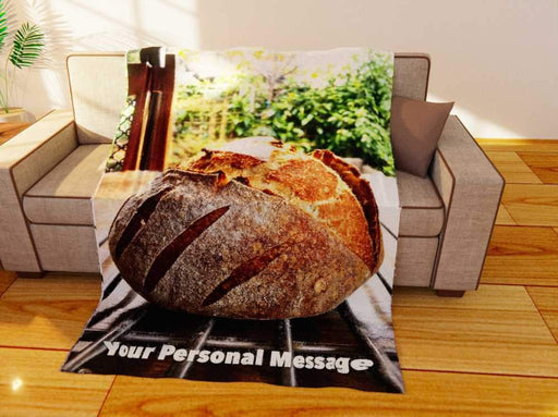 a fleece blanket flat on the couch, the blanket having an image of a sourdough loaf of bread sat upon a table in a kitchen, along with a printed personal message