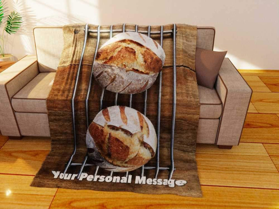 a fleece blanket flat on a couch, the blanket having an image of two sourdough loaved of bread sat on a metal wire tray, along with a printed personal message