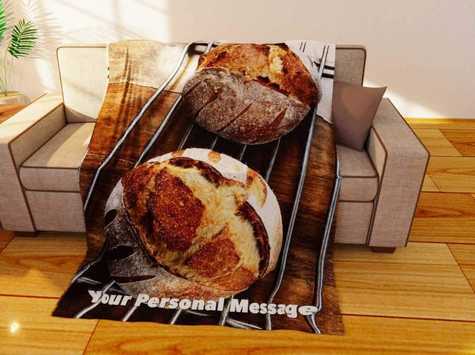 a fleece blanket flat on a couch, the blanket having an image of two sourdough loaved of bread in the centre sat on a metal tray, seen from an angle, along with a printed personal message