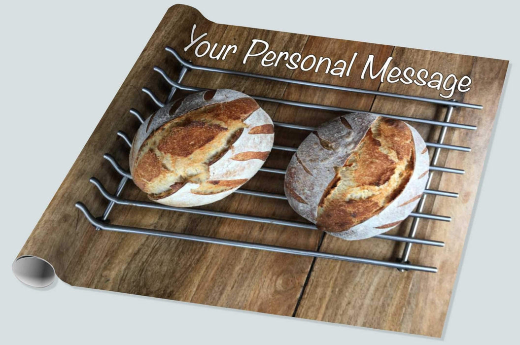 A roll of wrapping paper showing a pair of sourdough loaves resting on a metal tray upon a brown wooden table, along with a personal message