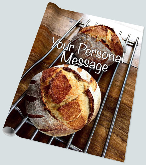 A roll of wrapping paper showing a pair of sourdough loaves resting on a metal tray upon a brown wooden table, along with a personal message, seen from an angled position
