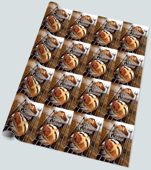 A roll of wrapping paper showing a pair of sourdough loaves resting on a metal tray upon a brown wooden table, along with a personal message, seen from an angled position, the image repeated in a mosaic pattern