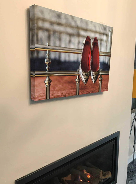 A canvas print of a pair of high heel stiletto shoes hung on the metal frame of a bed, as seen from the foot of the bed, the canvas print is hung above a fire place in a living room