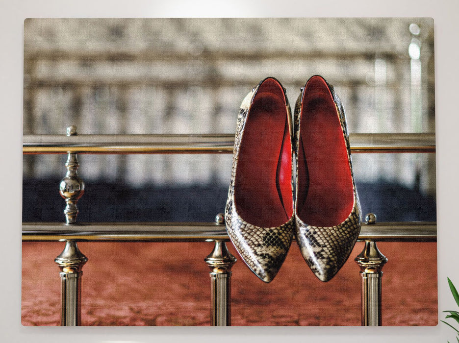 A pair of high heel stiletto shoes hung on the metal frame of a bed, as seen from the foot of the bed