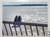 A pair of purple shoes sat on a blue metal fence on the shore of an ocean, with the ocean sparkling in the background