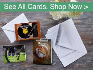 A series of greeting cards laid down on a grey wooden table, along with some envelopes, a pen and white flowers, there is a banner at the top saying See All Cards Shop Now, the banner is in white writing with green background