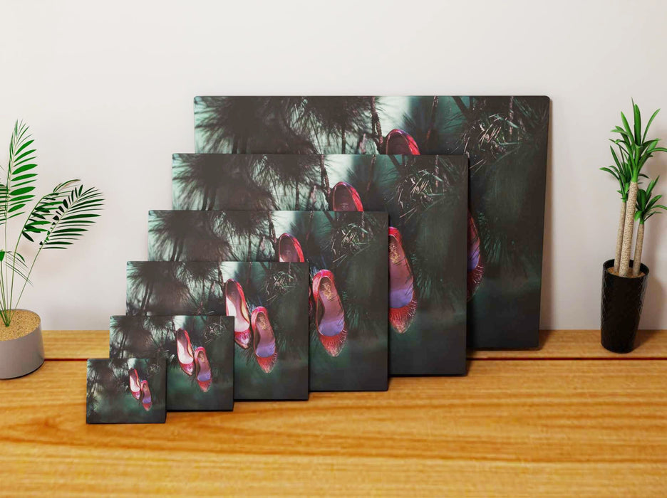 Multiple canvas prints of different sizes, each canvas print on a wall, the print showing a pair of red high heel shoes hanging from the branch of a tree, the prints are all leaning against each other against a wall