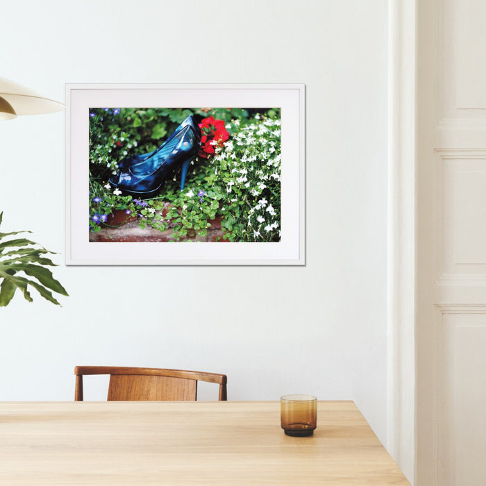 A framed print with a white frame hung in a kitchen setting, the print showing a pair of blue high heel shoes surrounded by flowers