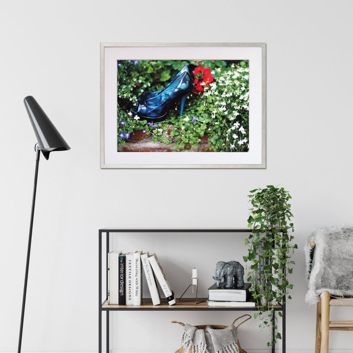 A framed print with a silver frame hung in a living room setting, the print showing a pair of blue high heel shoes surrounded by flowers