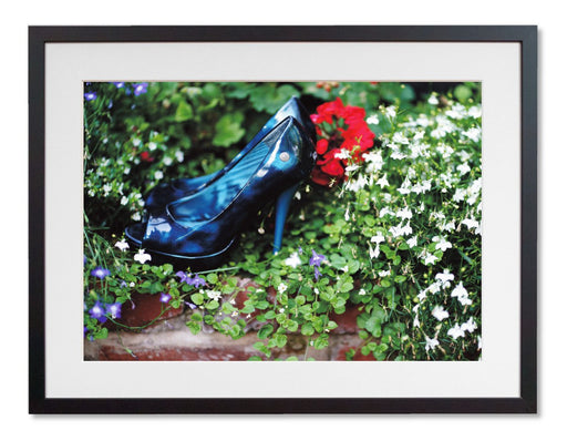 A framed print with a black frame, the print showing a pair of blue high heel shoes surrounded by flowers