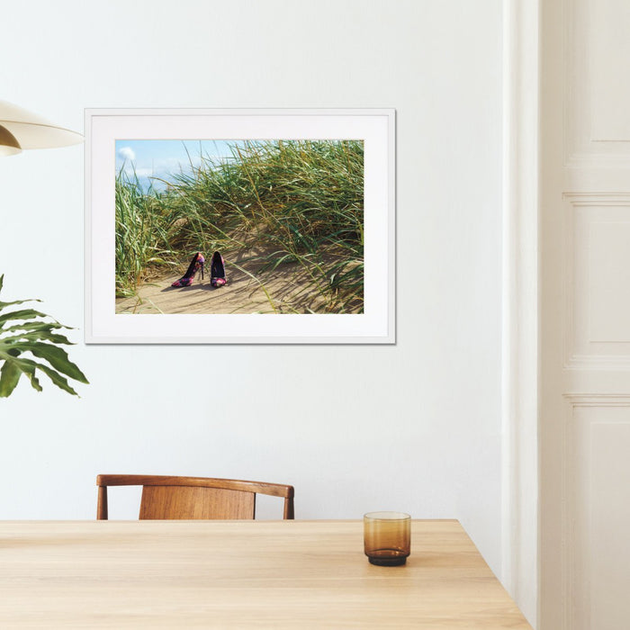 A framed print with a white frame on the walls of a kitchen, the print showing a pair of high heel shoes on the beach at the bottom of some sand hills