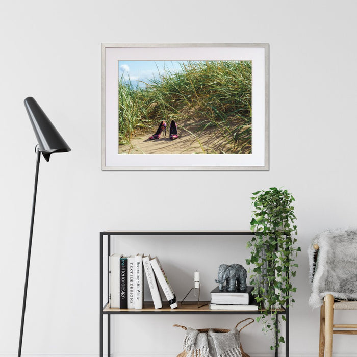 A framed print with a silver frame on the wall of a living room, the print showing a pair of high heel shoes on the beach at the bottom of some sand hills