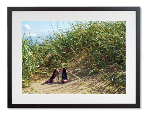 A framed print with a black frame, the print showing a pair of high heel shoes on the beach at the bottom of some sand hills