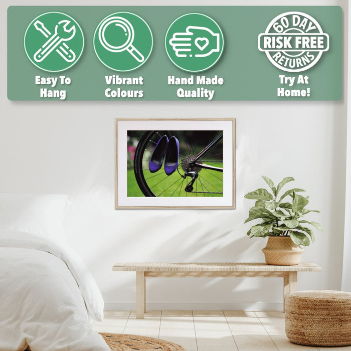 A framed print with a natural frame hung on the wall of a bedroom, the print showing a pair of purple high heel shoes hung off the spokes of the rear wheel of a road bike