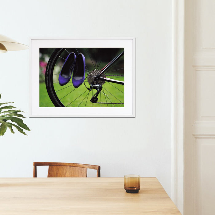 A framed print with a white frame on the wall of a kitchen, the print showing a pair of purple high heel shoes hung off the spokes of the rear wheel of a road bike