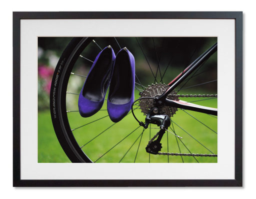 A framed print with a black frame, the print showing a pair of purple high heel shoes hung off the spokes of the rear wheel of a road bike