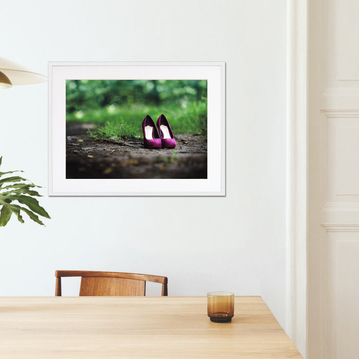 A framed print with a white frame hung on the walls of a kitchen, the print showing a pair of purple high heel shoes on a path through a woodland area