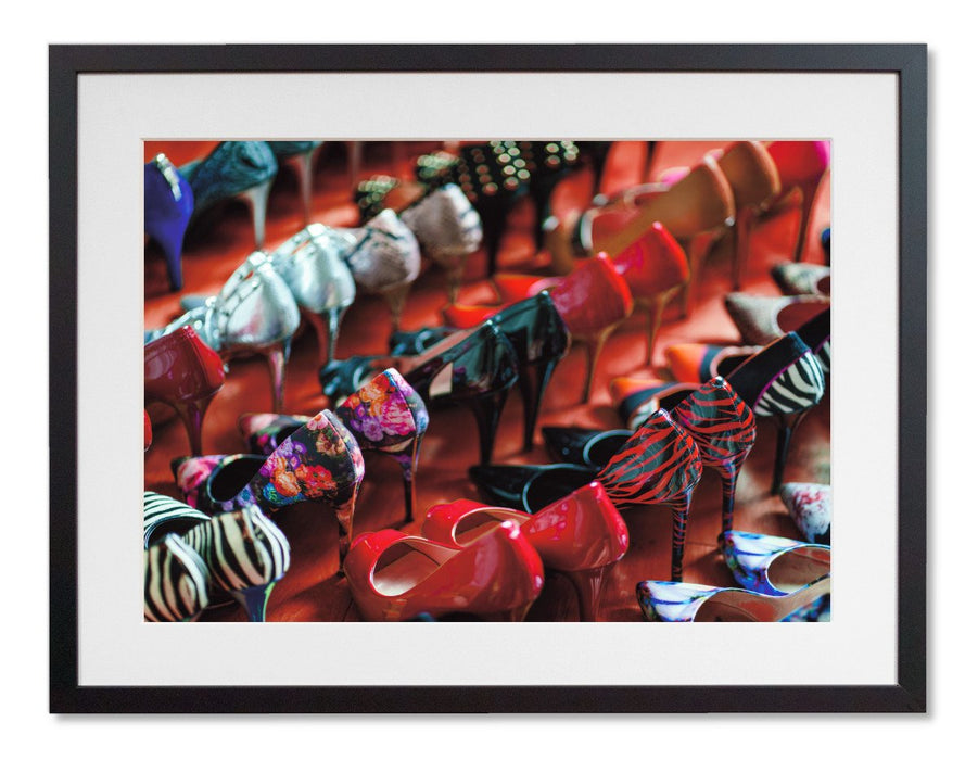 A framed print with a black frame, the print showing a multiple rows of high heel shoes all of different colours