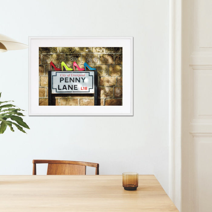 A framed print with a white frame hung on the wall of a kitchen, the print showing four coloured high heel shoes sat on top of the penny lane road sign in liverpool