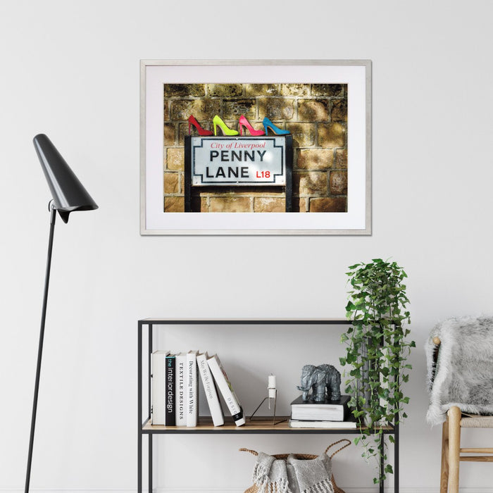 A framed print with a black frame hung on the wall of a living room, the print showing four coloured high heel shoes sat on top of the penny lane road sign in liverpool