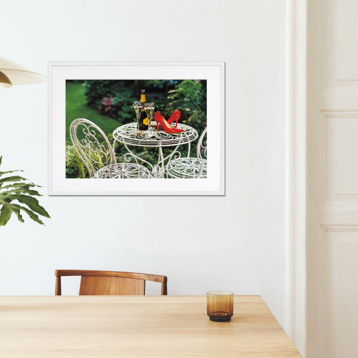 A framed print with a white frame hung on the wall of a kitchen, the print showing a pair of red high heels on a garden table next to a bottle of fizzy wine and two poured glasses