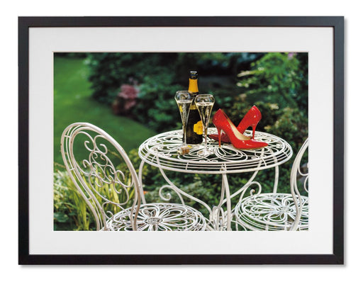 A framed print with a black frame, the print showing a pair of red high heels on a garden table next to a bottle of fizzy wine and two poured glasses