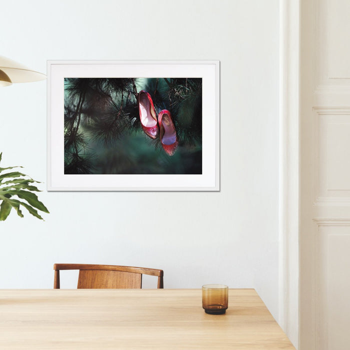 A framed print with a white frame hung on the wall of a kitchen, the print showing a pair of ruby high heel shoes hanging of the brances of a tree, so that it looks like the shoes are growing on the tree