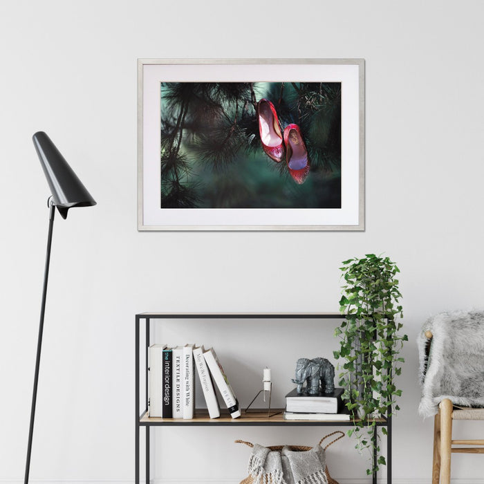 A framed print with a silver frame hung on the wall of a living room, the print showing a pair of ruby high heel shoes hanging of the brances of a tree, so that it looks like the shoes are growing on the tree