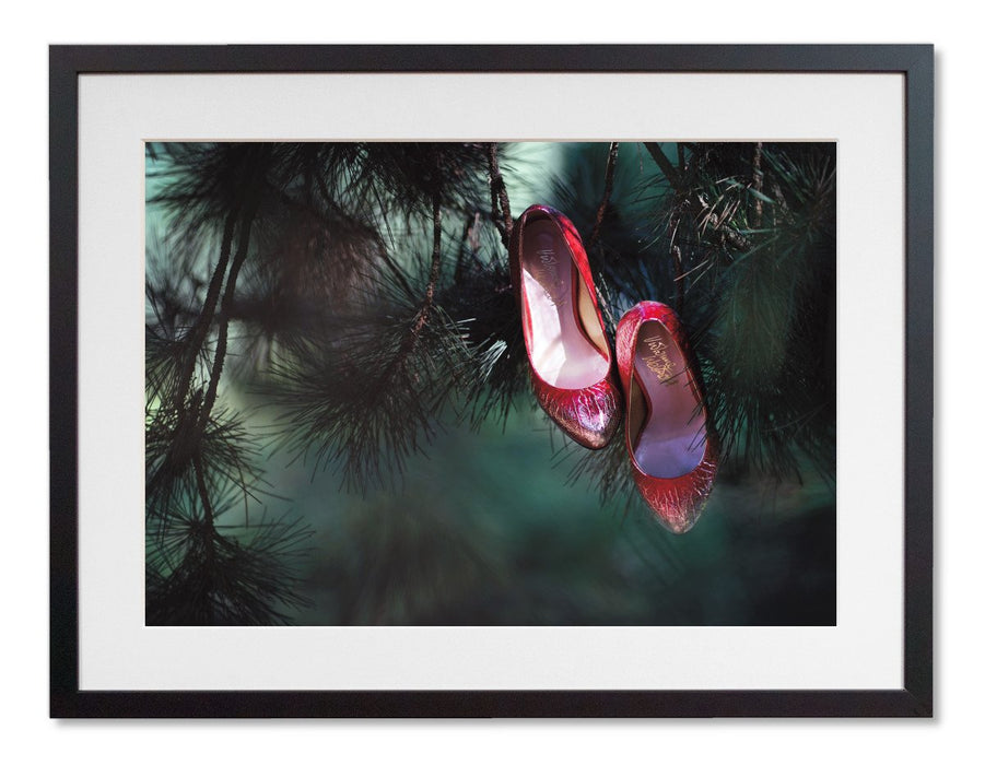 A framed print with a black frame, the print showing a pair of ruby high heel shoes hanging of the brances of a tree, so that it looks like the shoes are growing on the tree