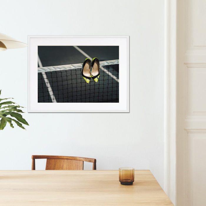 A framed print with a white frame hung on on the wall of a kitchen, the print showing pair of yellow and black high heel shoes hung over a tennis net on a tennis court