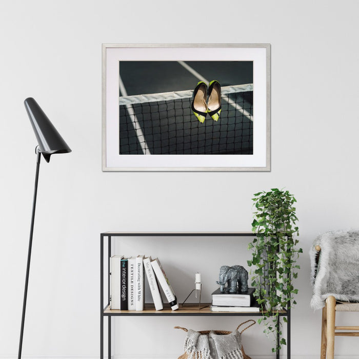 A framed print with a black frame hung on the wall of a living room, the print showing pair of yellow and black high heel shoes hung over a tennis net on a tennis court