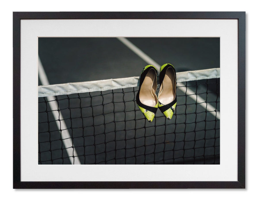 A framed print with a black frame, the print showing pair of yellow and black high heel shoes hung over a tennis net on a tennis court