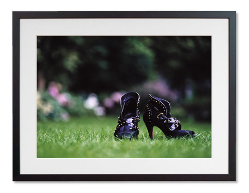 A framed print with a black frame, the print showing a pair of purple high heeled ankle boots sat on the grass in a garden