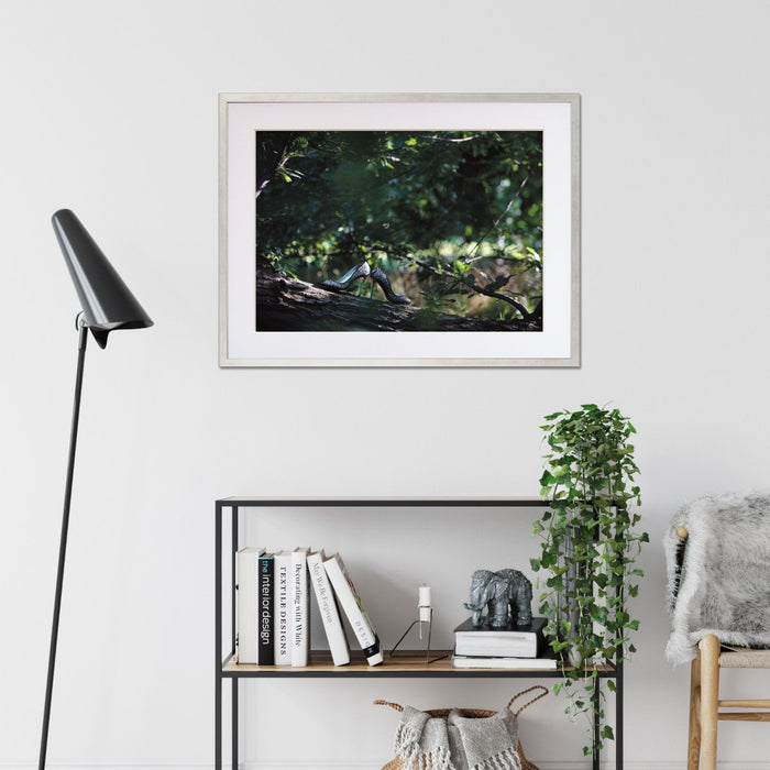 A framed print with a black frame hung on the walls of a living room, the print showing a pair of high heel shoes on the branch of a tree in a forest