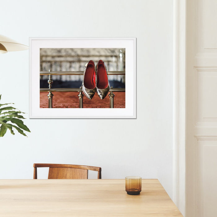 A framed print with a white frame hung on the wall of a kitchen, the print showing a pair of high heel shoes hung off the rails of a four poster bed