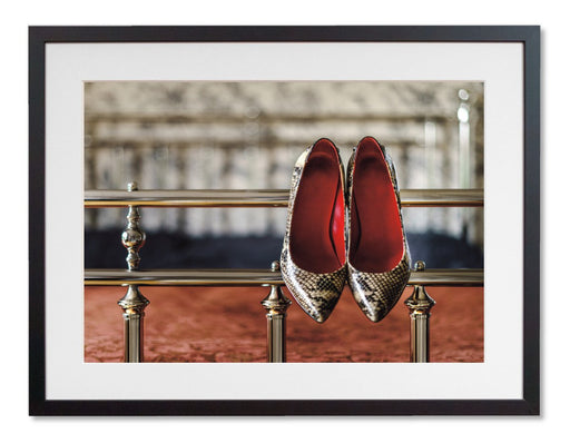 A framed print with a black frame, the print showing a pair of high heel shoes hung off the rails of a four poster bed